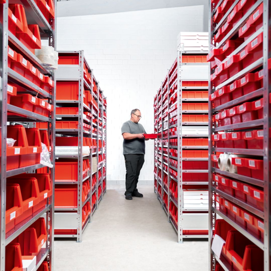 Legbordstellingen met rode magazijnbakken. In het midden staat en man in het grijsgekleed die een magazijnbak uit de stelling heeft gepakt en de hoge kwaliteit inspecteert van SCHULTE stellingen.